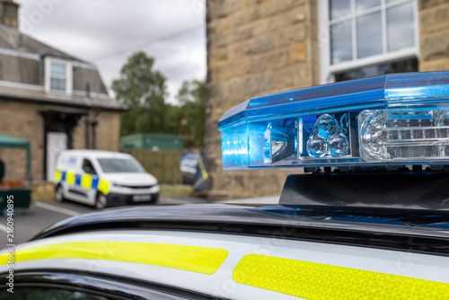 Close up of a blue emergency light bar mounted on the roof of a British police vehicle with high visibility yellow markings and another marked police van softly blurred in the background