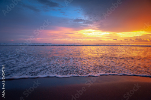 Beautiful sunset over a calm tropical ocean, with gentle waves rolling toward a sandy beach and warm golden sunlight reflecting across the water. The dramatic sky transitions from deep blue and purple
