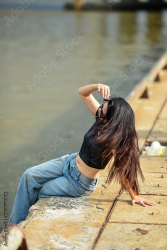 Young woman with long dark hair posing on a concrete pier by the water in sunglasses and denim jeans