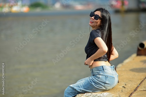Smiling Asian woman in sunglasses, black crop top and blue jeans sitting on concrete riverside embankment