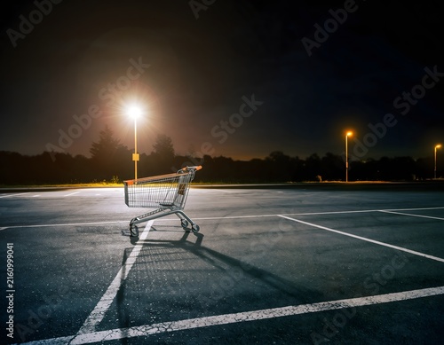 Abandoned shopping cart in an empty parking lot at night under streetlights.