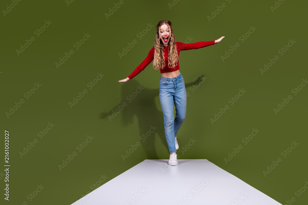 Naklejka premium Young woman in red top and jeans walks a white runway against green background with excited smile