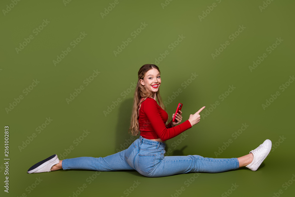 Naklejka premium Young woman performing a full split in red top jeans and white sneakers holding a phone smiling against a green background