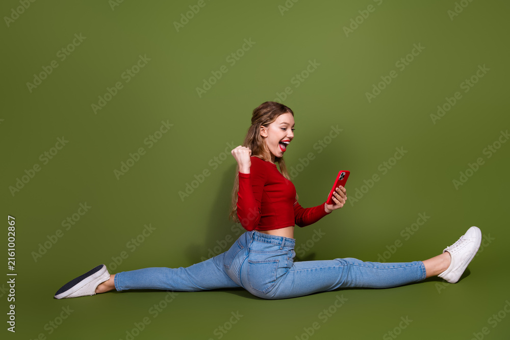 Naklejka premium Young woman performs split on green background while using red smartphone joyfully
