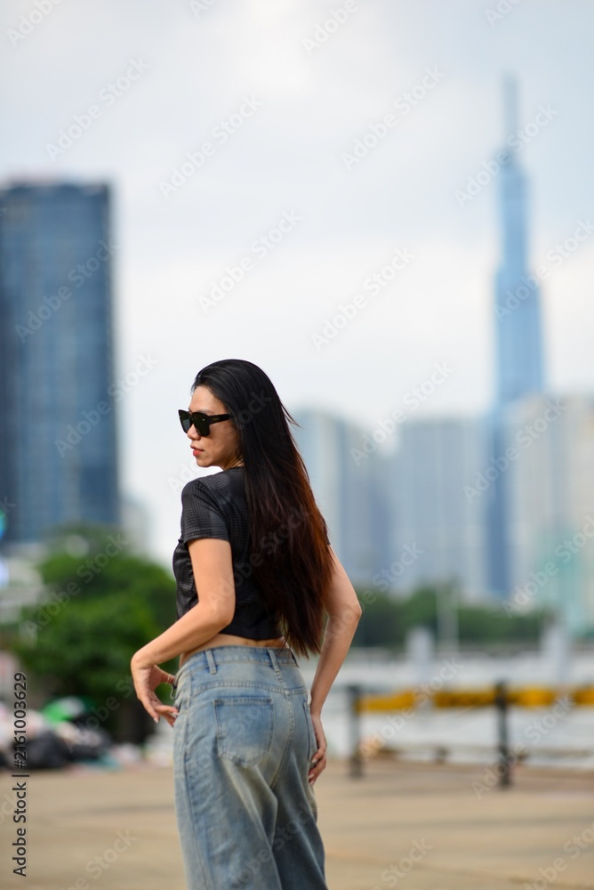 Fototapeta premium Stylish young Asian woman in black crop top and baggy jeans posing outdoors with modern city skyline in background