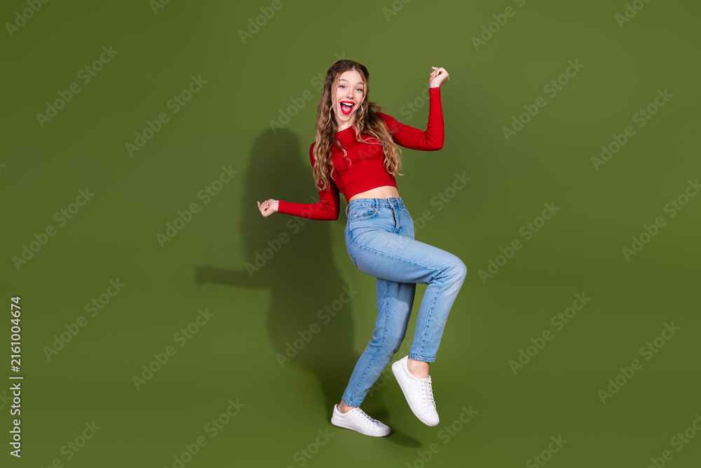 Naklejka premium Young woman jumps with joy in red top and jeans against green studio background for lifestyle fashion advertising use