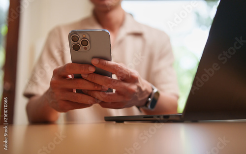Foto Man using smartphone beside laptop at home, representing mobile browsing, online communication, messaging, digital lifestyle, remote work, internet connectivity, multitasking, and modern technology