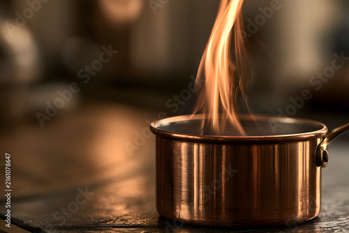 Vibrant flames rise from a rustic copper pot, illuminating a kitchen setting with a warm, inviting glow. A dynamic scene of culinary artistry.