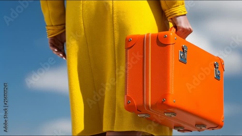 A person in a bright yellow coat holds a vintage orange suitcase against a blue sky background.