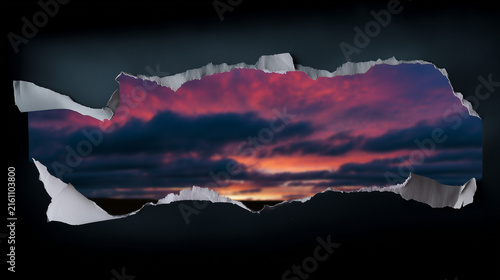 A breathtaking sunset sky with vibrant red, orange, and purple clouds, viewed through a jagged hole torn in dark paper, symbolizing hope, hidden beauty, and looking beyond.