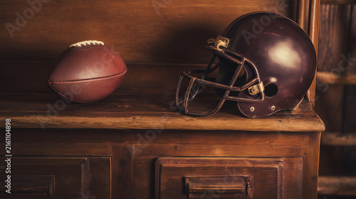 A vintage, aged American football helmet sits beside a classic leather football on an old, dark wooden cupboard, capturing the essence of historical sports memorabilia and nostalgic gridiron memories.