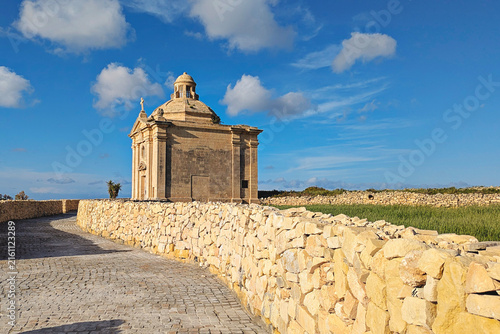 Kappella San Nikolaw chapel beside paved path and traditional Maltese stone walls, green fields and bright blue sky with scattered clouds in rural Malta