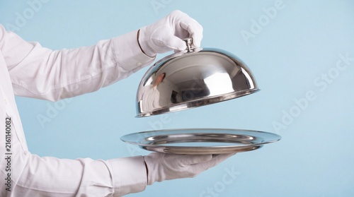 Waiter wearing white gloves lifting polished silver cloche from empty round serving tray isolated background with serving platter against light blue background