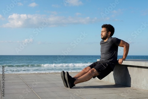 Man performing tricep dips on bench near ocean during outdoor calisthenics workout. Bodyweight fitness and strength training concept
