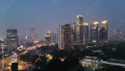 Aerial drone view of Jakarta's Sudirman business district at night featuring illuminated skyscrapers, glowing office buildings, vibrant city lights, and heavy traffic after office hours, showcasing th
