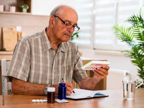 Senior man with eyeglasses writing notes while organizing medicine in pill organizer and preparing his daily dose at home.