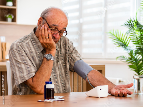 Senior man sitting in his living room and measuring blood pressure at home while feeling worried about his health and cardiovascular problems.