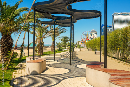 Photography Contemporary black metal pergola of curved shape creating patterned shadows on pavement in a seaside resort park with palms and bamboo under a blue sky