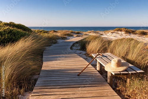 peaceful wooden boardwalk through coastal sand dunes leading to the atlantic ocean with rustic bench, straw hat and walking stick in warm golden sunlight