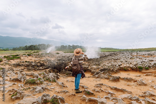 Cuadro en lienzo Rear view of hiker wearing hat and carrying backpack explores rocky landscape where steam rises from ground