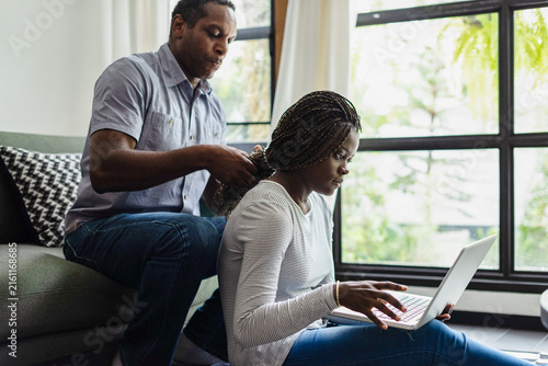 Canvas Print Man braiding woman's hair as she uses a laptop at home.
