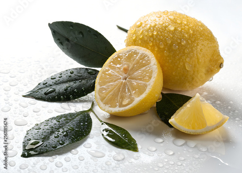Fresh whole lemon sliced half wedge dark green leaves covered in glistening water droplets on white surface