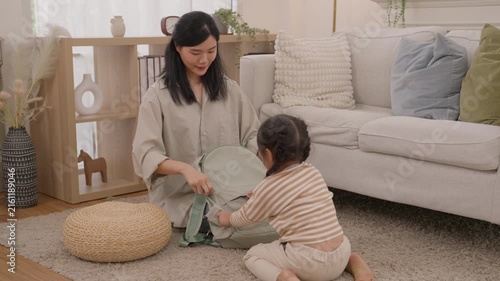 Mother and child packing backpack while Asian parent and toddler play indoors cozy living room, sitting beside fabric basket and sofa, creating warm caring family moment with natural light and home