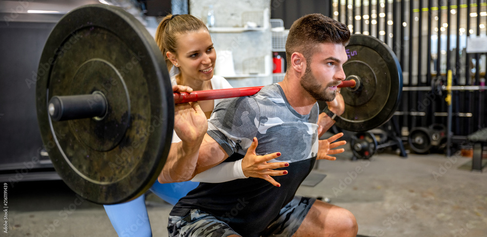 Fototapeta premium Female personal trainer spotting male athlete during barbell squat exercise in gym.