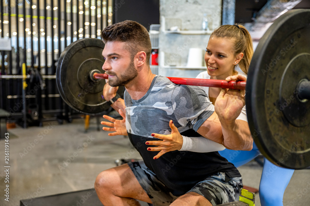 Fototapeta premium Female fitness coach assisting male client with barbell back squat training in modern gym.