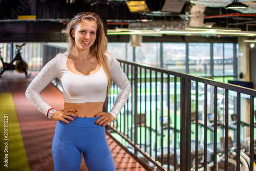 Smiling fit woman standing near balcony railing posing confidently in modern gym.