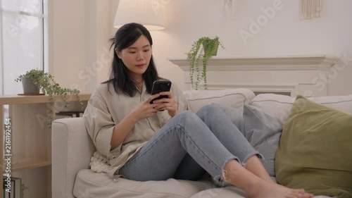 Asian woman using smartphone on sofa in bright home interior with houseplant, cushion, natural light, lifestyle relaxation, modern comfort, quiet indoor leisure and calm living room atmosphere