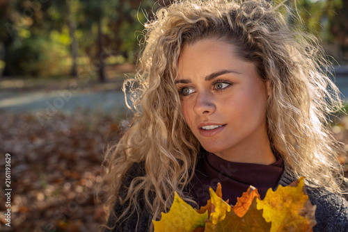 Closeup shot of natural looking beautiful young blonde with curly hair holding dry autumn leaves at sunset 