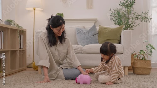 Asian mother and toddler family play with pink toy piggy bank carpet cozy living room home, enjoying warm indoor bonding, savings activity, joyful learning, and meaningful parent child connection
