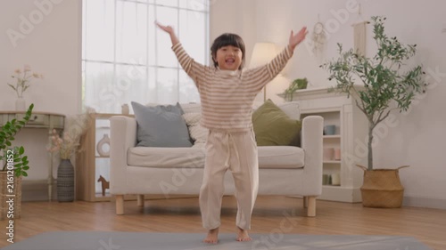 Child Girl Dancing Bright Living Room Home, Smiling Striped Shirt and Beige Pants, Indoor Portrait with Outstretched Arms Near Comfortable Sofa and Indoor Plant, Joyful Energy Asian Family Lifestyle