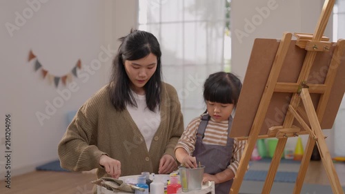 Asian woman and child painting together beside easel in colorful home art studio, creative education activity with vibrant supplies, focused family bonding and joyful artistic expression indoors
