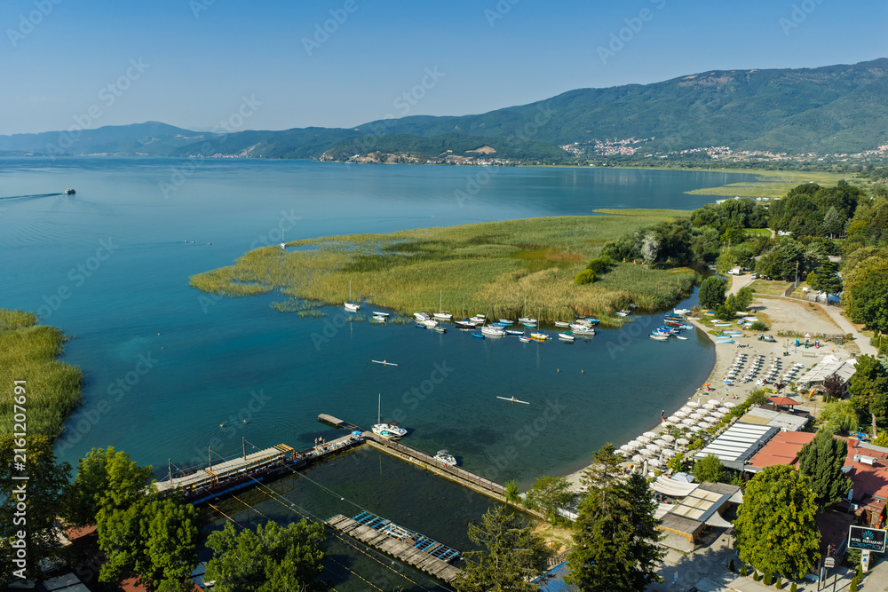 Fototapeta premium Aerial drone view of Struga in North Macedonia with Black Drin River flowing into crystal clear Lake Ohrid, boats, sunny summer landscape, waterfront buildings and scenic city panorama