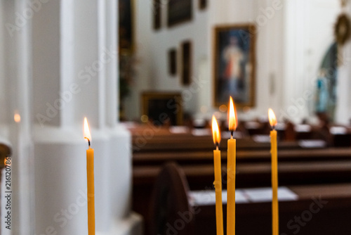 Canvas Print Yellow wax candles burning with bright flames inside a dimly lit church, capturing a peaceful, spiritual, and meditative atmosphere with blurred church pews in the background