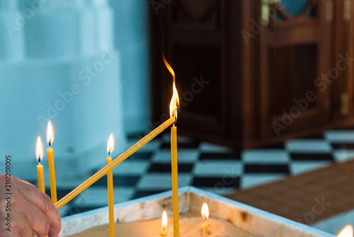 A close-up view of a person's hand using an existing flame to light a thin yellow church candle in a sand box inside a church with a checkered floor background.