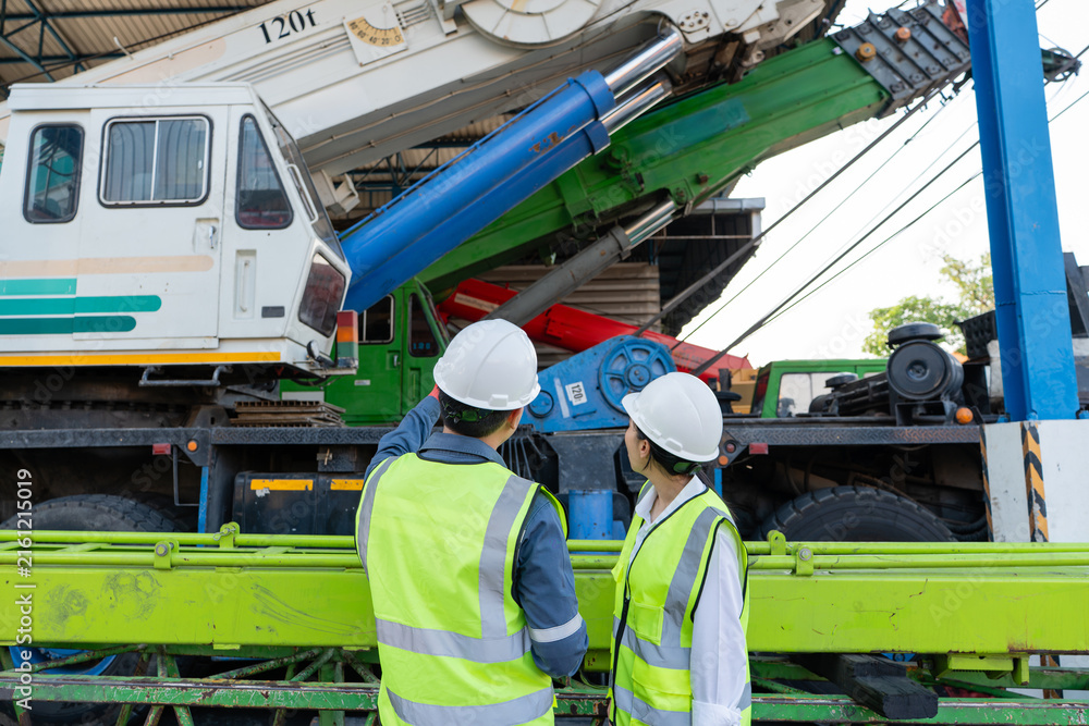 Fototapeta premium Performing a crane preventive maintenance inspection, two engineers wearing hard hats examine the hydraulic cylinder and boom