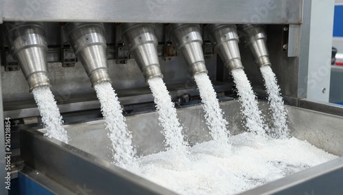 White plastic polymer pellets pouring from an industrial machine into a large storage container in a manufacturing facility.
