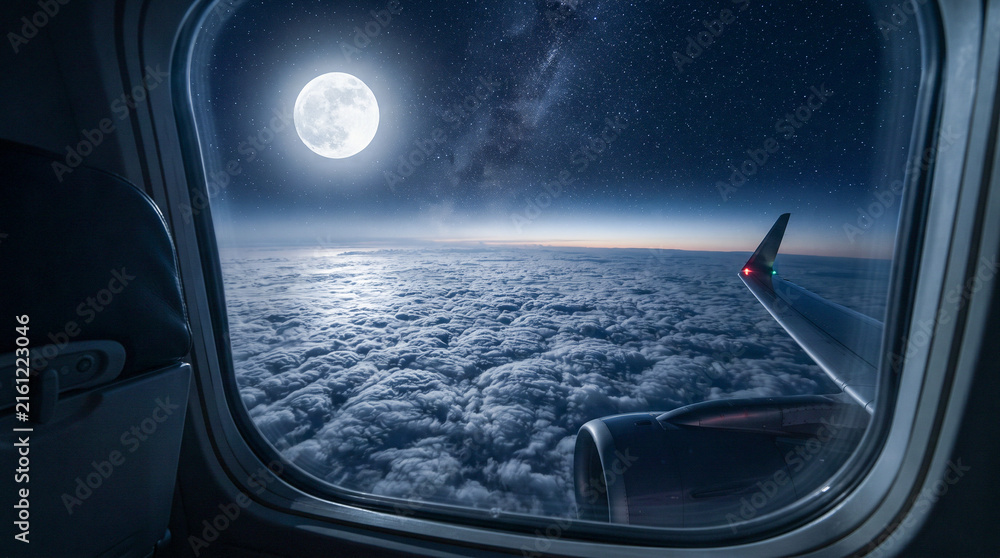 Naklejka premium Vista nocturna desde la ventanilla de un avión mostrando la luna llena, la Vía Láctea y un mar de nubes iluminado sobre el horizonte.