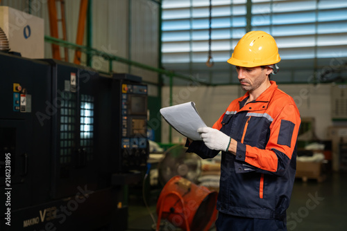Male technician wearing yellow safety helmet reading inspection document near CNC machine in a factory, quality control, industrial engineering and manufacturing
