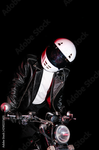 Tired male motorcyclist in white helmet and leather jacket resting on ground next to motorcycle wheel at dark night. Road trip fatigue and night travel concept