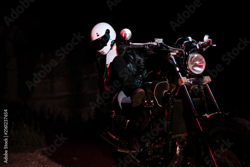 Tired male motorcyclist in white helmet and leather jacket resting on ground next to motorcycle wheel at dark night. Road trip fatigue and night travel concept