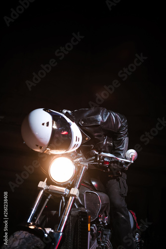 Tired male motorcyclist in white helmet and leather jacket resting on ground next to motorcycle wheel at dark night. Road trip fatigue and night travel concept
