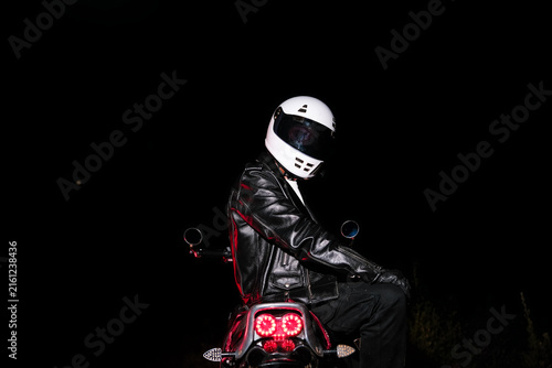 Low key portrait of motorcyclist in white helmet and black leather jacket sitting on motorcycle in dark night. Night ride, dramatic lighting and biker lifestyle concept