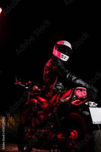 Low key portrait of motorcyclist in white helmet and black leather jacket sitting on motorcycle in dark night. Night ride, dramatic lighting and biker lifestyle concept