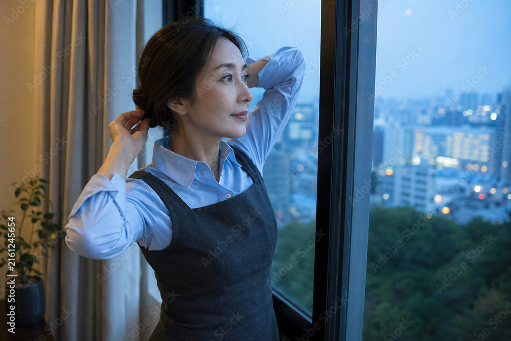 Fototapeta premium 夜の窓辺で青いシャツのエプロン姿で髪を整える女性