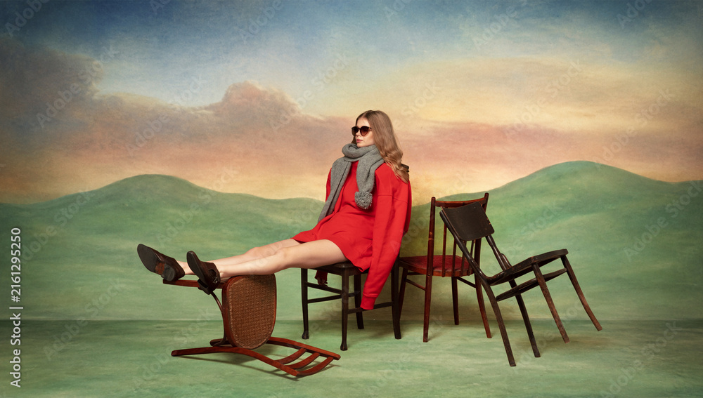 Fototapeta premium Woman in vivid red coat poses among scattered chairs across softly painted theatrical dream landscape. Concept of luxury lookbooks, magazine covers and fashion ads.