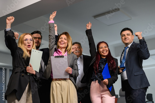 Group of happy people celebrating success in office setting during daytime with papers in hand and smiles on their faces after reaching a goal or completing a task